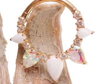 May include: Gold-toned septum ring featuring a row of clear, sparkling crystals and pear-shaped opalescent stones. The jewelry is set against a neutral background, highlighting the intricate design and iridescent accents.