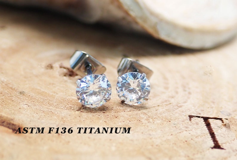 May include: A pair of silver titanium stud earrings with clear round cubic zirconia stones. The earrings are on a wooden surface with the text "ASTM F136 TITANIUM" visible.