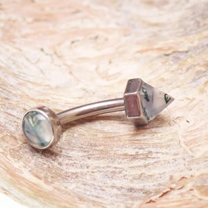 May include: A curved belly button ring with a silver-colored metal bar. One end features a round, light green stone, while the other has a geometric, triangular-shaped stone with green inclusions.