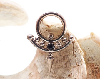 May include: A silver-colored septum ring with a hinged closure. The design features a curved row of small silver balls, a central black stone, and a dangling silver cone. The ring is displayed on a piece of light-colored wood.