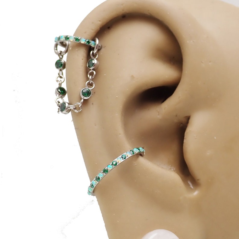 Puede incluir: Un par de piercings de cart&iacute;lago de oreja con piedras preciosas verdes y blancas. El piercing superior es una cadena con piedras preciosas verdes y el piercing inferior es un aro con piedras preciosas verdes y blancas.