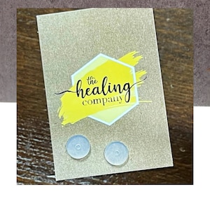May include: A pair of small, clear, round, adhesive earplugs on a brown card with the text "the healing company" in black lettering on a yellow hexagon with a yellow paint stroke behind it.