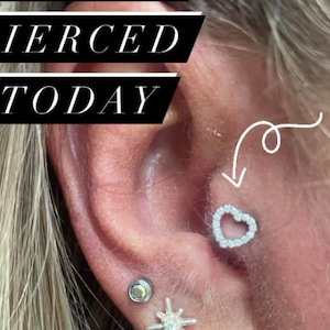 Könnte beinhalten: Nahaufnahme eines Ohres mit einem neuen Piercing. Das Piercing ist ein kleiner, silberner Herzring mit klaren Steinen am Rand. Der Text "PIERCED TODAY" ist in der oberen linken Ecke des Bildes sichtbar.
