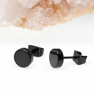 May include: A pair of black stud earrings featuring a circular, flat-faced design with a polished finish. Each earring includes a thin post and a secure butterfly-style backing, presenting a simple, geometric aesthetic with a solid black color.