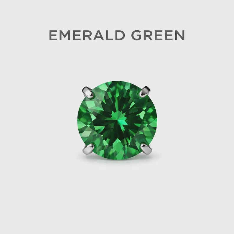 May include: A single, round emerald green gemstone with a faceted surface, set in a silver or white metal setting. The text "EMERALD GREEN" is displayed above the gemstone against a white background.