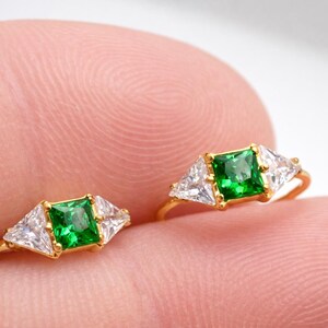 May include: Two gold rings, each featuring a square emerald-cut green gemstone flanked by two triangular white gemstones. The rings are set on a gold band.