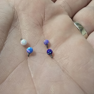 Pode incluir: Quatro pequenos brincos de joalharia corporal iridescentes semelhantes a opalas. Os brincos têm formato de bola e são em tons de branco, azul e roxo. Cada brinco tem um pequeno pino roscado para inserção. A joalharia é exibida em tom de pele claro.