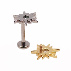 May include: Two pieces of body jewelry. One is a silver-colored labret stud with a starburst design. The other is a gold-colored starburst top. Both have a central hole.