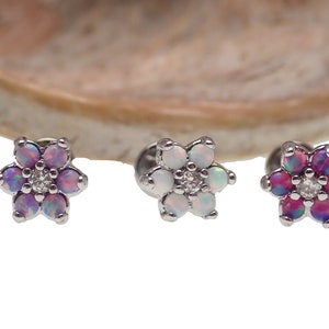 Pode incluir: Três peças de joalheria corporal de prata em forma de flor com pedras de opala. As flores têm uma pedra central e seis pedras circundantes. As pedras são em tons de rosa, branco e roxo.