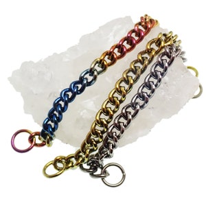 May include: Three colorful chain bracelets with a curb link design. The bracelets are in shades of blue, gold, and silver, with a rainbow-colored ring at the end of the blue bracelet. The chains are displayed on a white, crystalline surface.