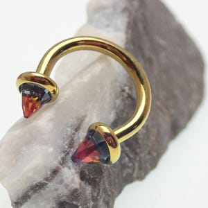 Puede incluir: Un piercing dorado en forma de herradura con gemas cónicas multicolores en cada extremo. Las gemas hacen una transición de naranja a morado. La joya está colocada sobre un fondo neutro y texturizado.