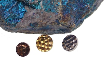 May include: Three small, round metal objects with textured surfaces. One is bronze, one is gold, and one is silver. The objects are displayed against a white background with a large, colorful mineral formation in the background.