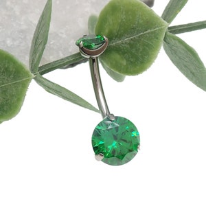 May include: A silver belly button ring with two green gem stones. The larger gem stone is round and the smaller gem stone is oval shaped.