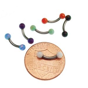 May include: Six curved barbell body jewelry pieces with colorful opal-style spherical ends. One piece rests on a copper United States one cent coin for scale, while five others are arranged around it in blue, purple, green, orange, and black.