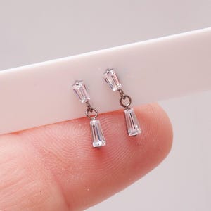 May include: A pair of silver-toned dangle earrings. Each earring features a small, rectangular, clear crystal at the top, connected by a small loop to a larger, rectangular crystal pendant. The earrings are displayed against a light background.