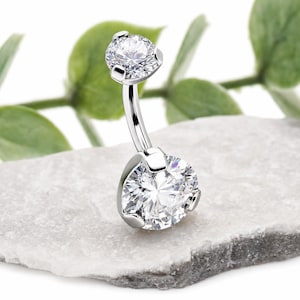 May include: A silver-toned curved barbell navel ring featuring two clear, round-cut faceted stones held in place by four-prong settings. The larger stone is positioned at the base, while the smaller stone sits at the top of the curved metal bar.