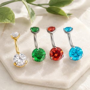 May include: Four curved barbell belly button rings with round faceted gemstones. One gold-tone ring features a clear stone, while three silver-tone rings feature green, red, and bright blue stones respectively, each with a smaller matching top gem.
