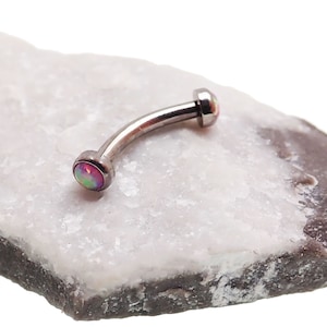 May include: A curved barbell body jewelry piece featuring a metallic silver-toned shaft with two circular ends, each set with a round, iridescent pink opal-like stone that displays flashes of green and orange light across its smooth surface.