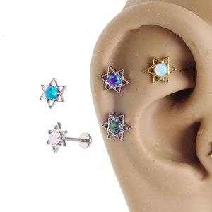 May include: Three silver Star of David body jewelry pieces with opal gemstones. The jewelry is shown on a model's ear.