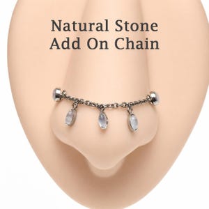 May include: A nose chain with natural stone pendants. The chain is silver-colored and connects to the nose with two small, round studs. Three oval-shaped stones dangle from the chain, with the text "Natural Stone Add On Chain" above.