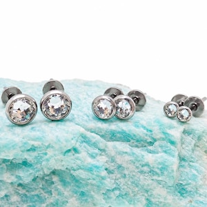 May include: Three pairs of stud earrings featuring clear, round-cut faceted stones set in polished silver-toned metal bezels. Each earring has a flat, circular backing post. The earrings are arranged in descending order of size from left to right.