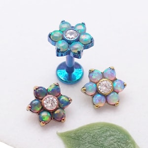 May include: Three flower-shaped body jewelry pieces. Two are flat-backed, with a central clear gem surrounded by opalescent stones. The third is a post with a flower top, featuring similar opalescent stones and a central gem. The jewelry has gold and blue tones.