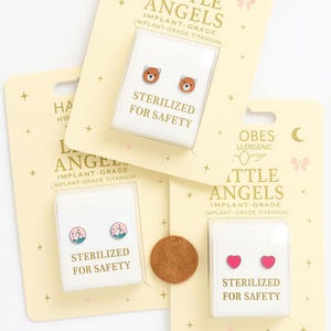 Puede incluir: Se muestran tres pares de pendientes hipoalergénicos. Un par presenta diseños de osos, otro tiene forma de donut y el tercero muestra corazones rosas. Cada par está empaquetado con el texto "STERILIZED FOR SAFETY".