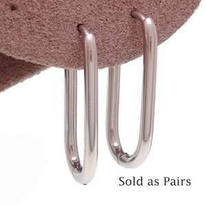 May include: A pair of polished rose gold-toned metal hoop earrings with an elongated oval shape. The earrings are attached to a textured, light brown suede-like material. The text "Sold as Pairs" is printed in black at the bottom right corner.