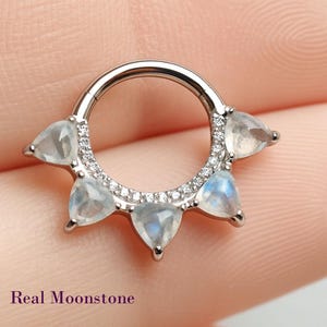 May include: A silver-colored septum ring with five pear-shaped, translucent moonstone gems. The ring is adorned with small, clear crystals and the text "Real Moonstone" is visible at the bottom.