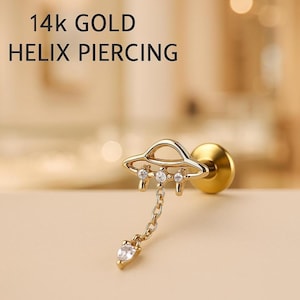 May include: A 14k gold helix piercing featuring a small, detailed UFO design. The UFO has three clear, round gemstones and a dangling teardrop-shaped gem. The text "14k GOLD HELIX PIERCING" is displayed in black.