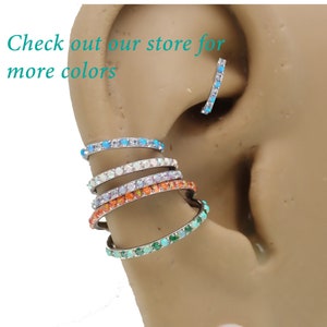 Puede incluir: Primer plano de una variedad de anillos de cart&iacute;lago de oreja con gemas de colores. Los anillos est&aacute;n apilados uno encima del otro, con el anillo inferior de color verde claro, el siguiente naranja, el siguiente blanco y el superior azul claro. Los anillos son todos de metal y tienen un acabado brillante. El texto "Check out our store for more colors" es visible en la imagen.