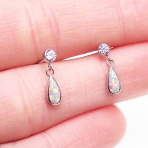 May include: A pair of silver-toned stud earrings featuring a round clear stone at the top and a dangling teardrop-shaped opal stone below. The opal displays iridescent flashes of color, and each earring is secured with a small metal jump ring.