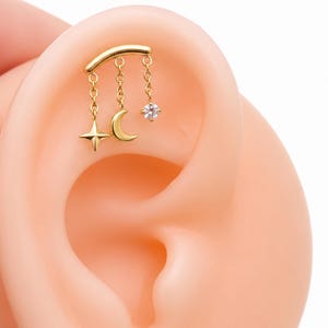 May include: A gold-tone ear cartilage earring featuring a curved bar with three dangling charms: a four-pointed star, a crescent moon, and a small round clear gemstone. The jewelry is positioned on the upper cartilage of a pale ear model.