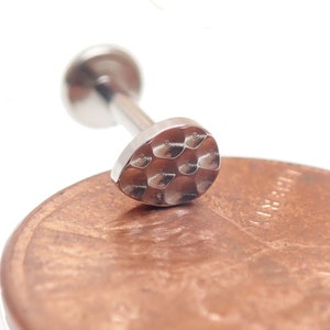 Puede incluir: Joyería corporal de plata con una parte superior redonda, plana y texturizada. La joyería está sobre un centavo.