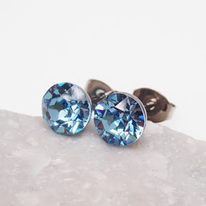 May include: A pair of round stud earrings featuring faceted light blue crystals. Each circular crystal is set in a metallic base with a post back, displaying a reflective, multi-faceted surface that catches the light with deep blue and silver tones.