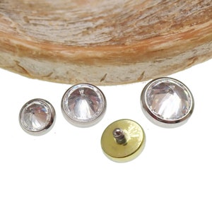 Op de afbeelding: Drie zilveren lichaamsieraden met heldere kristallen accenten. Eén stuk is een platte schijf met een schroefpost, de andere twee zijn rond met een platte achterkant.