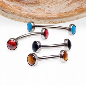 May include: Four curved barbell body jewelry pieces with metallic silver shafts. Each end features a smooth, round cabochon stone in different colors: bright blue, deep red, solid black, and a mottled brown tiger eye pattern.