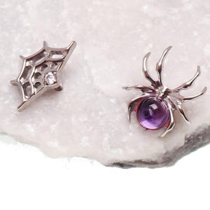 May include: A pair of silver-toned earrings consisting of one spider-shaped stud featuring a round purple cabochon stone and one web-shaped stud featuring a small clear round gemstone set within the metallic frame of the spider web design.