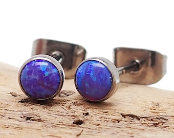 Könnte beinhalten: Ein Paar Ohrstecker mit runden, blau-violetten Opalsteinen, die in einem silberfarbenen Metall gefasst sind. Die Ohrringe werden auf einem Holzstück vor weißem Hintergrund präsentiert.