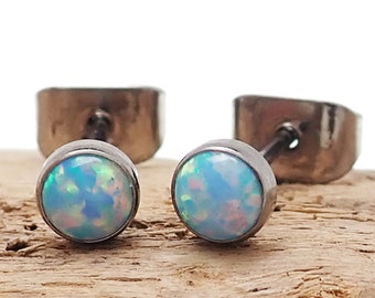 Könnte beinhalten: Ein Paar runde Ohrstecker mit einem hellblauen, opalartigen Stein, der in einem silberfarbenen Metall gefasst ist. Die Ohrringe werden auf einem Holzstück präsentiert. Die Ohrstecker und -verschlüsse sind ebenfalls sichtbar.