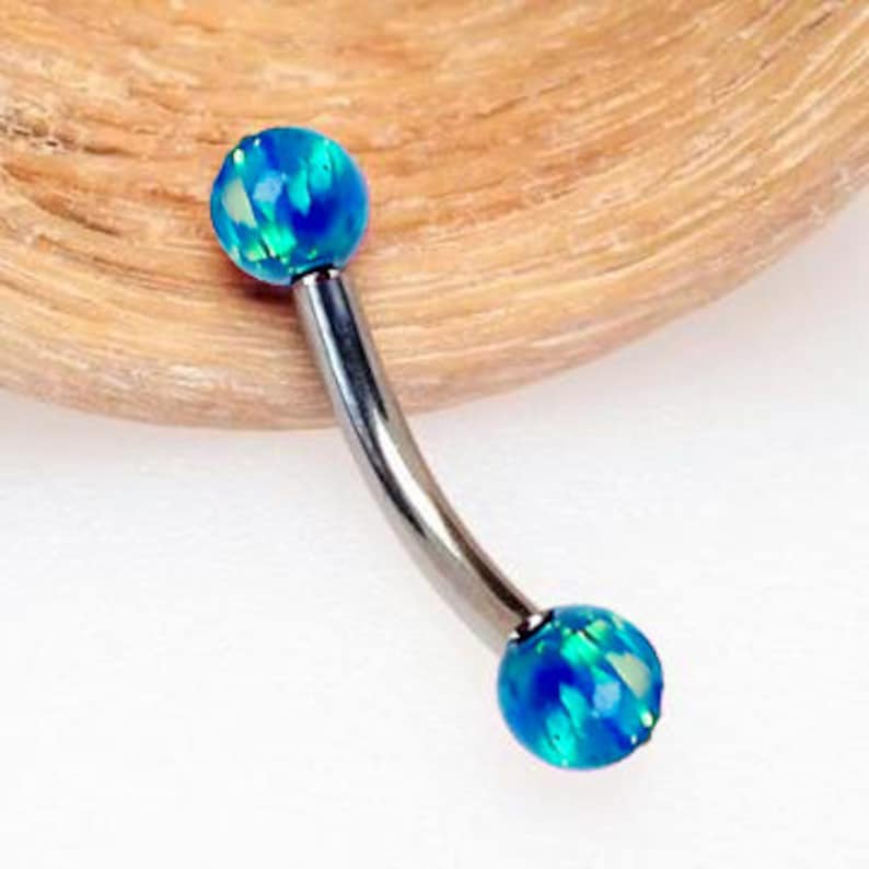 May include: A curved barbell body jewelry piece featuring two spherical ends made of blue opal with iridescent green and light blue flecks. The central curved bar is made of a smooth, polished silver-toned metal connecting the two opal spheres.