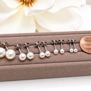 May include: Five pairs of white pearl stud earrings with silver-toned posts arranged in descending order of size on a brown velvet display tray, positioned next to a United States one-cent copper coin for scale reference.