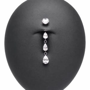 May include: A silver-toned belly button ring featuring a round clear gemstone at the top and a dangling vertical chain of three pear-shaped clear gemstones. The jewelry is displayed on a smooth, dark, oval-shaped surface with a central indentation.