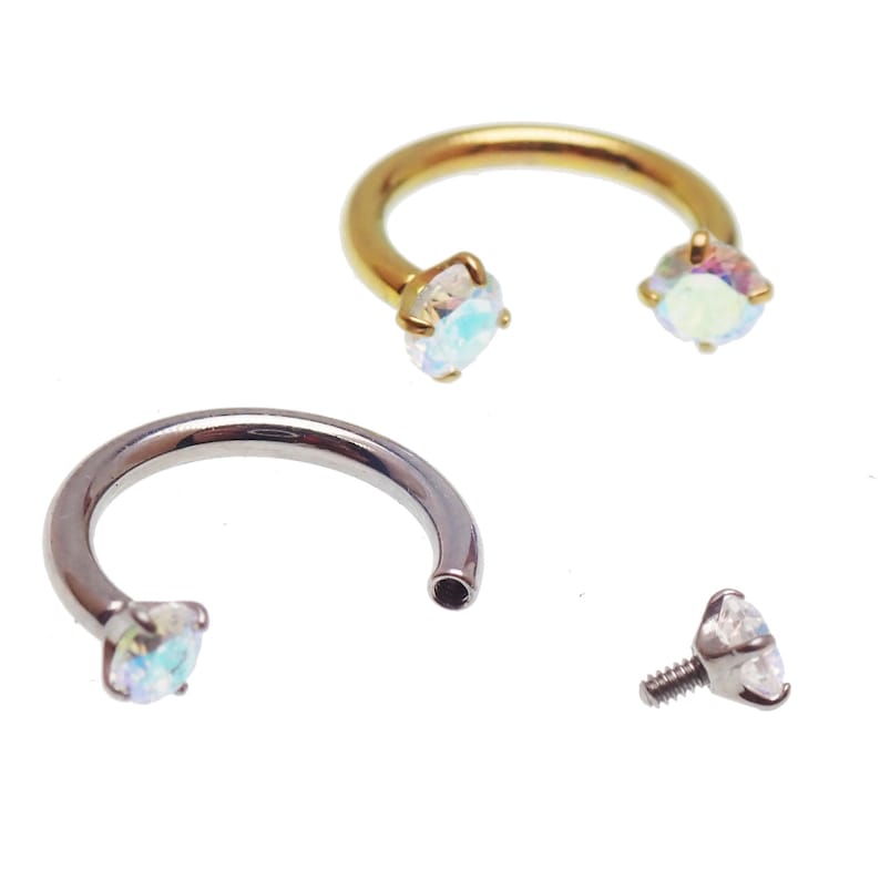 May include: A selection of body jewellery, including two horseshoe-shaped rings and a threaded stud. One ring is gold-toned, the other is silver-toned, each with a clear, iridescent gem at each end. The stud has a similar gem.
