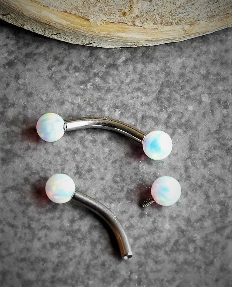 May include: Two curved barbell body jewelry pieces made of polished silver-tone metal. Each end features a smooth, round, iridescent white opal-like bead. One barbell is fully assembled, while the other is shown with one bead detached, revealing the screw.