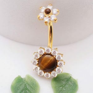 May include: A gold-colored belly button ring with a floral design. The top and bottom flowers have a brown center stone surrounded by clear, round gemstones. The curved bar is gold.