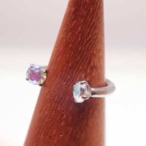 May include: Open-style ring featuring two iridescent, round gemstones. The silver-toned ring has gemstones that display pink, purple, and blue hues. The ring is displayed on a wooden cone.