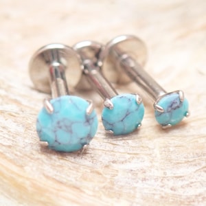 May include: Three silver-toned lip piercing studs with turquoise-colored, round stones. The stones have a marbled appearance with dark blue veining. The studs are displayed on a light-colored, textured surface.