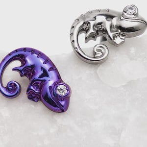 May include: Two chameleon-shaped jewelry pieces, one in a polished silver tone and the other in a metallic purple finish. Each lizard figure features a curled tail and a single clear, round-cut gemstone set into the eye area of the head.