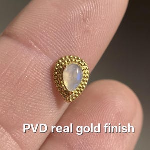 May include: A gold-colored, teardrop-shaped stud earring with a pearlescent, light-colored stone. The earring is surrounded by a textured, dotted border. The text "PVD real gold finish" is visible.